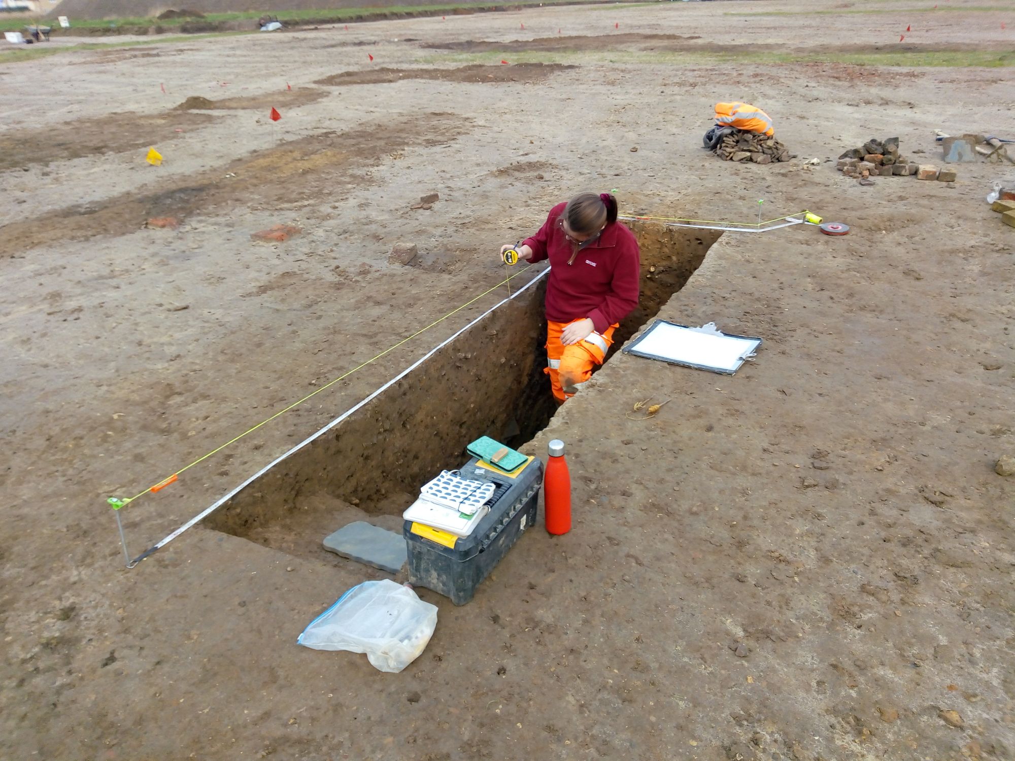 An archaeologist holds a measuring tape to record the section of a narrow dug slot.