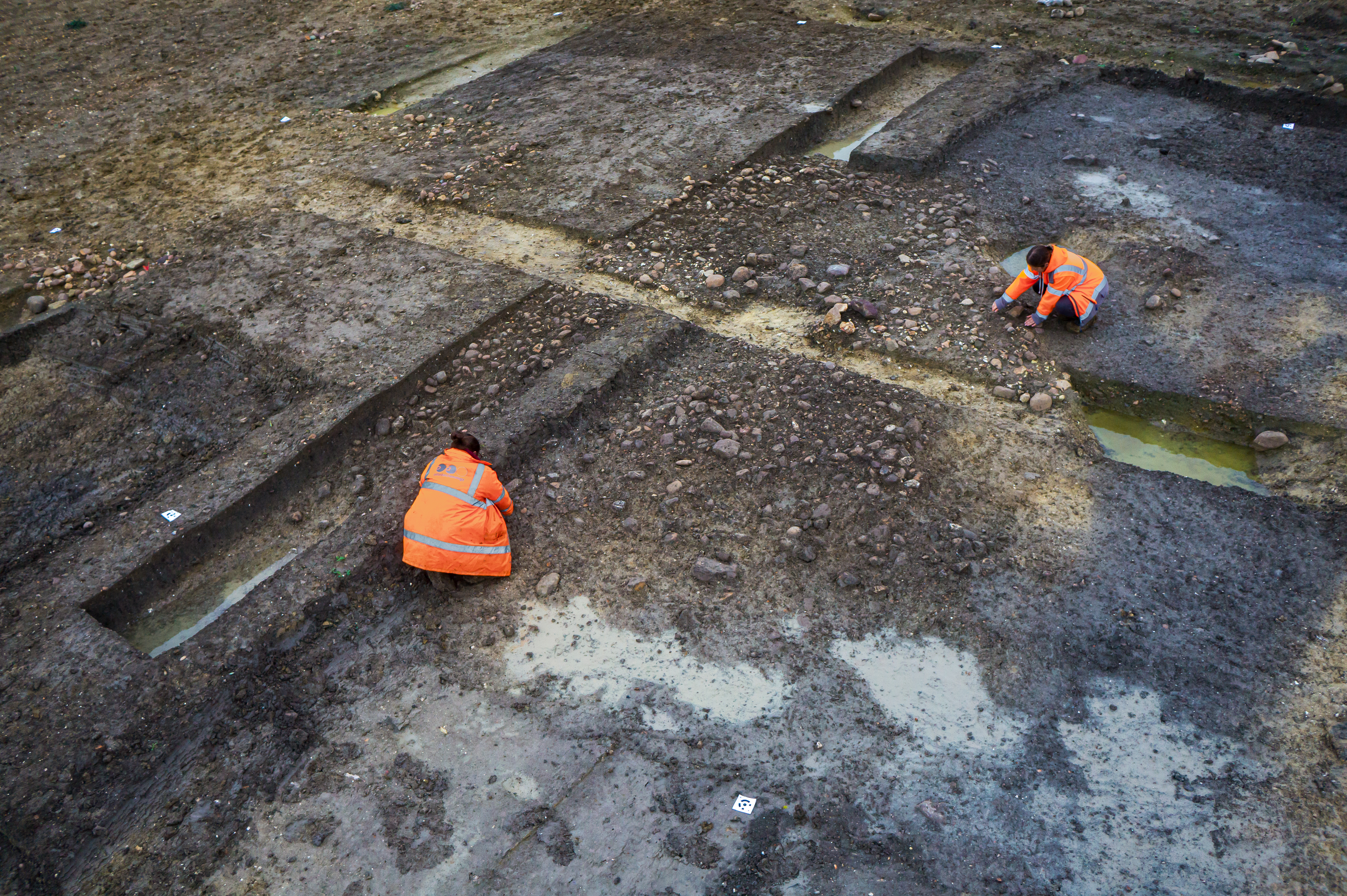 An aerial view of two archaeologists in high visibility clothing, cleaning an exposed area of deposited stones within a layer of dark soil.