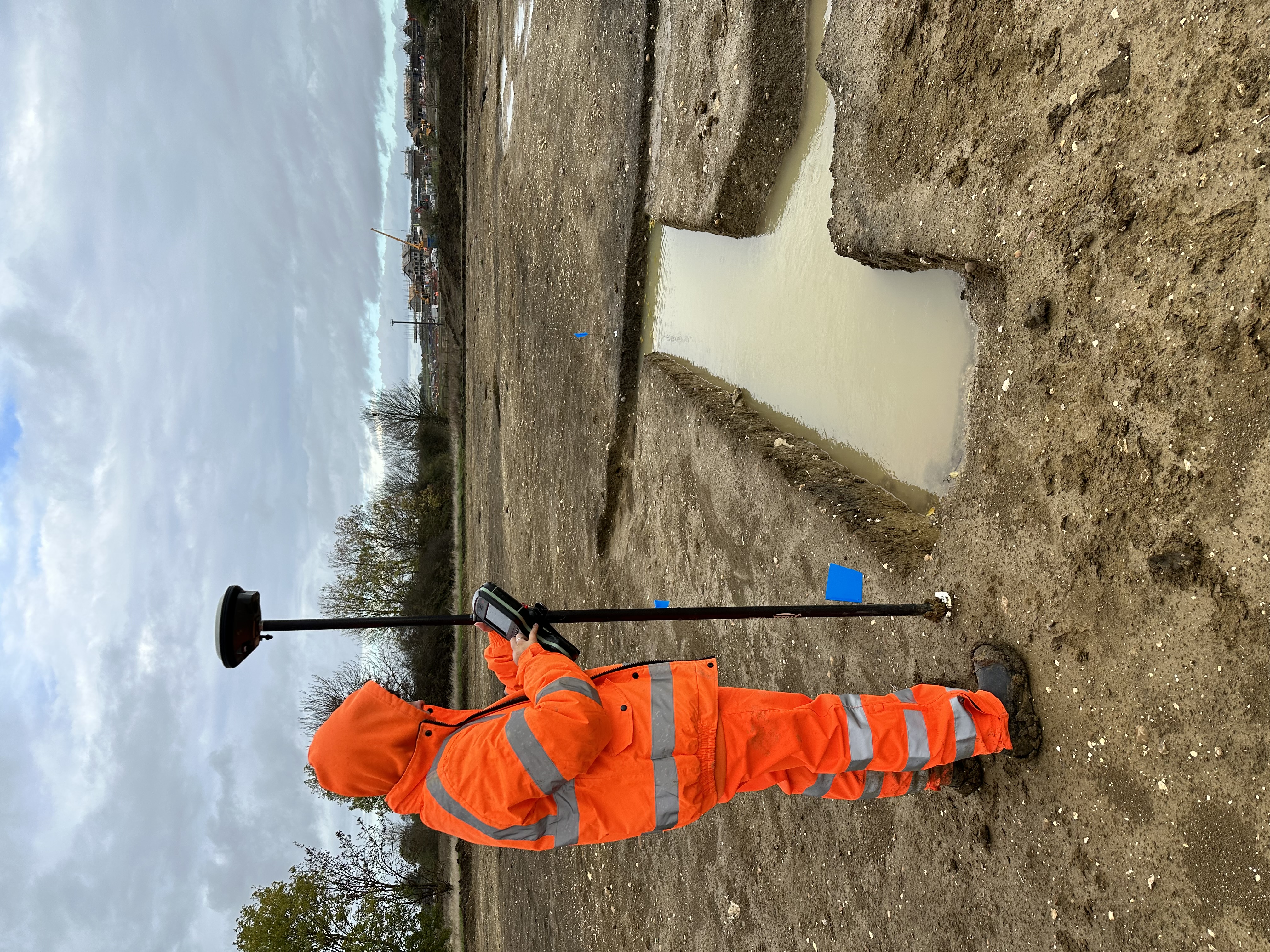 An archaeologist in high visibility clothing uses a tall GPS pole device to mark out the location of an excavated section that is filled with water
