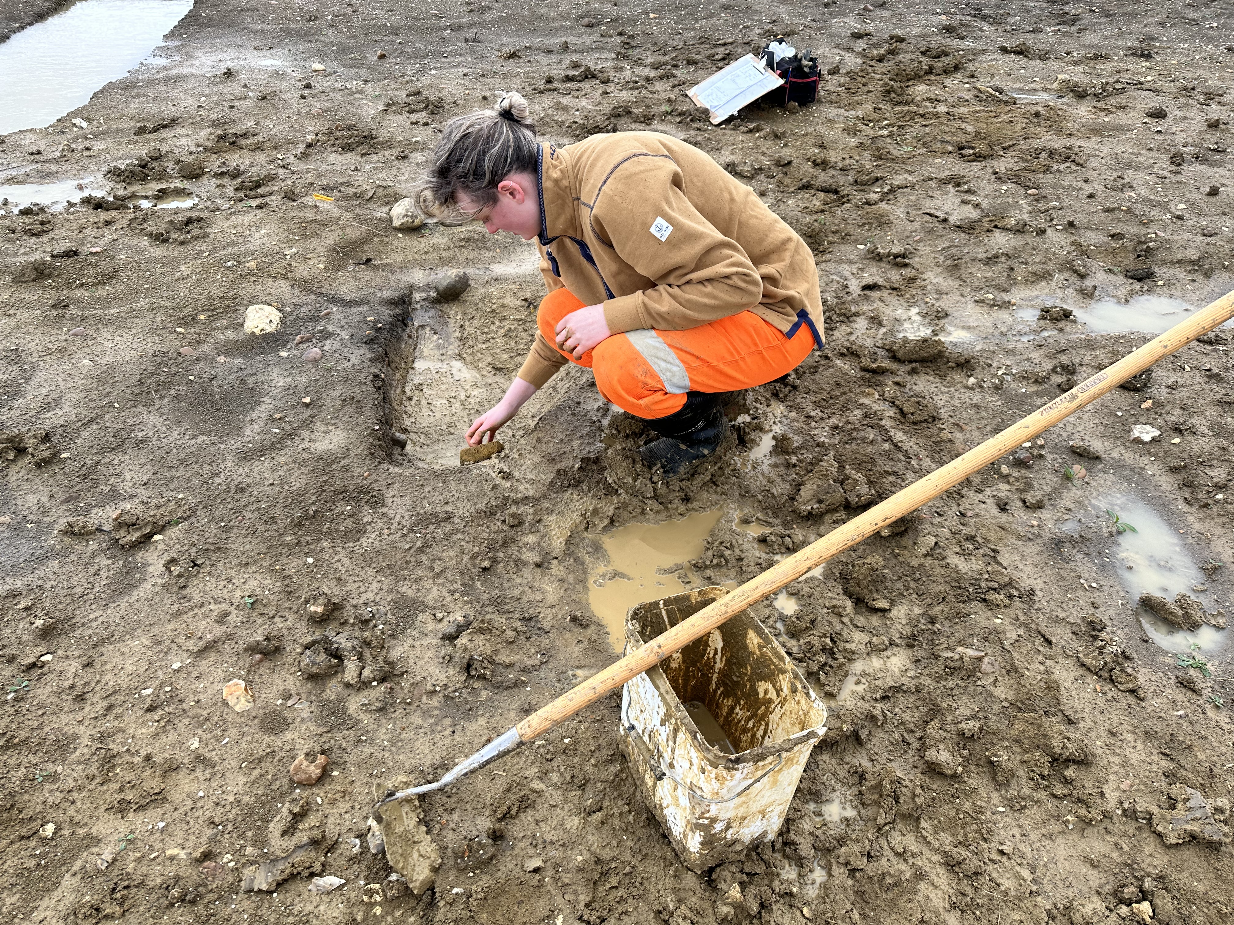 Graduate trainee archaeologist Ashley wearing high visibility orange trousers, surrounded by mud and puddles, uses a sponge to clear water from the bottom of her feature to get it ready for recording.