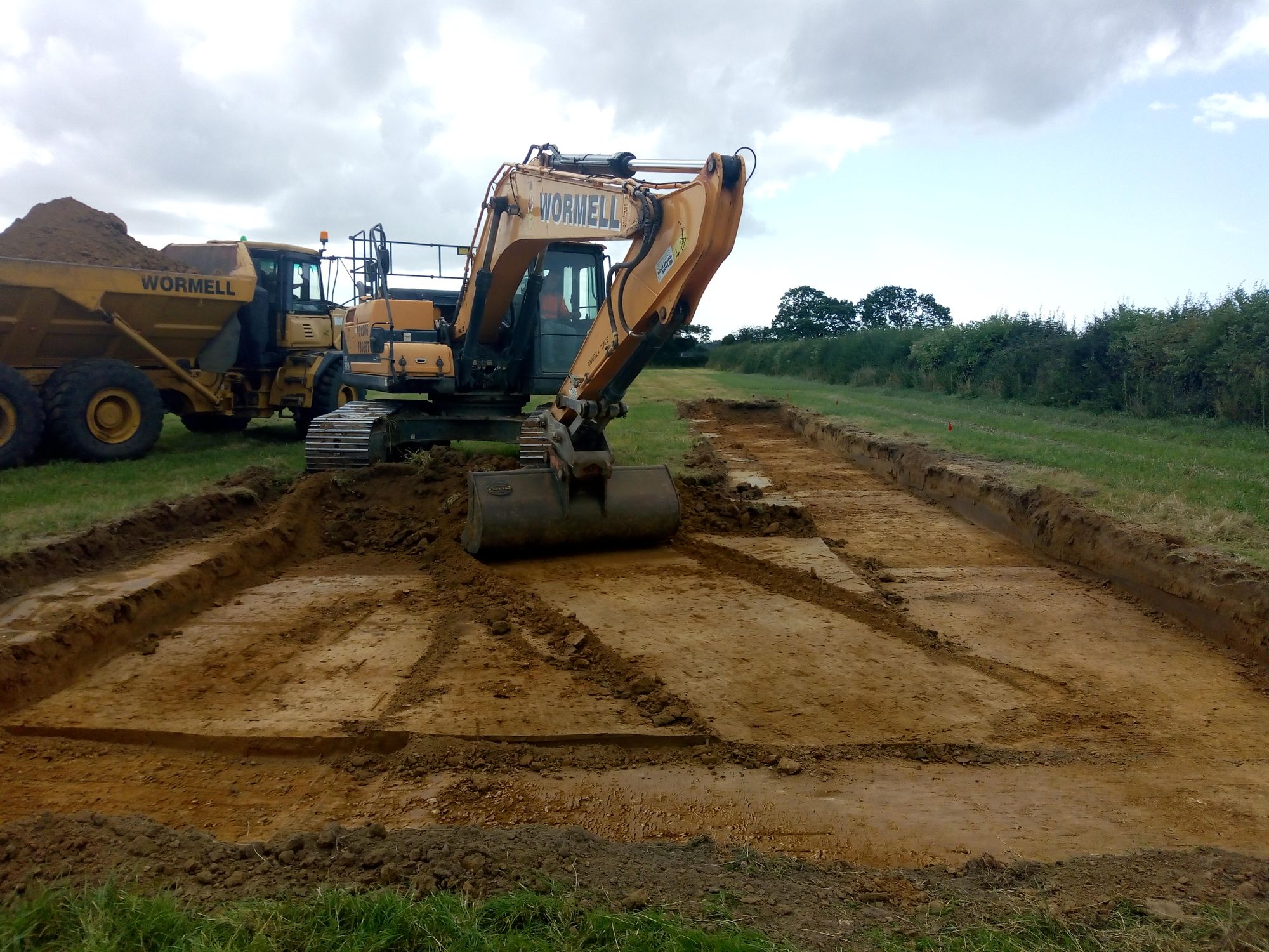 A digger arm peels back the subsoil from the excavation area, with a dumper truck behind.
