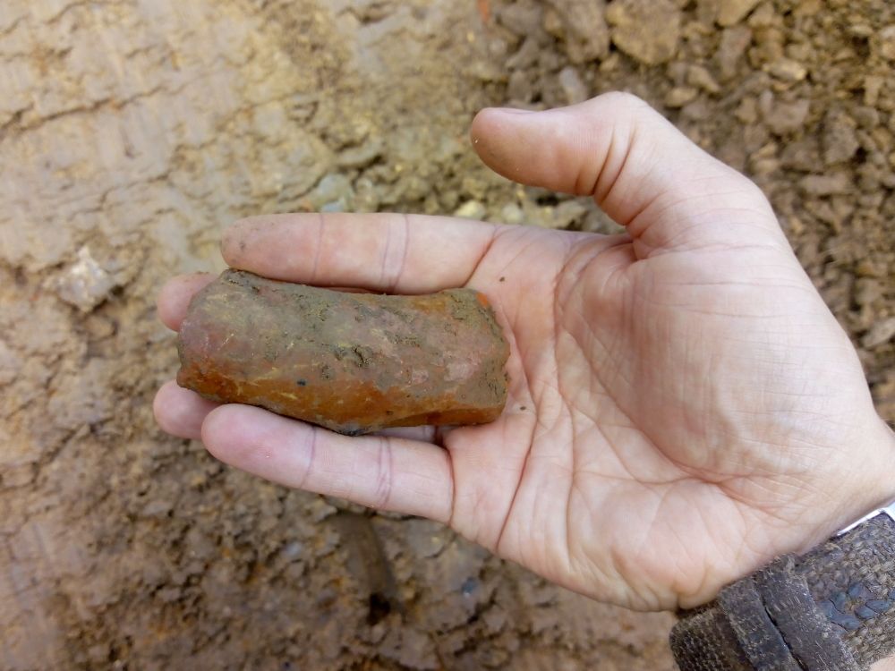 A hand holds a muddy fragment of orange-red glazed pottery.
