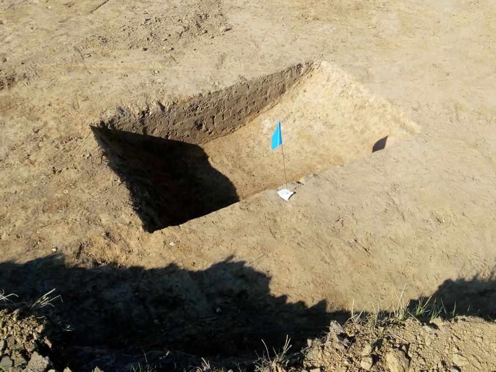 A hole dug in the ground to reveal the size and shape of a ditch, marked by a flag and tag.