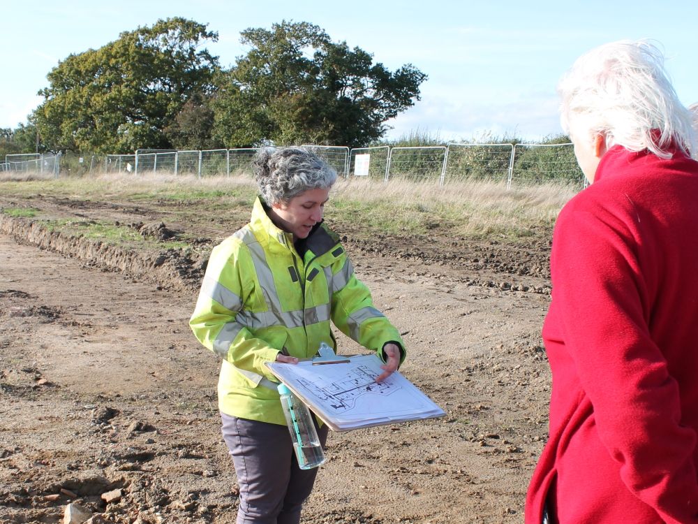 An archaeologist holds a clipboard with plan and points to what it shows during a site tour.