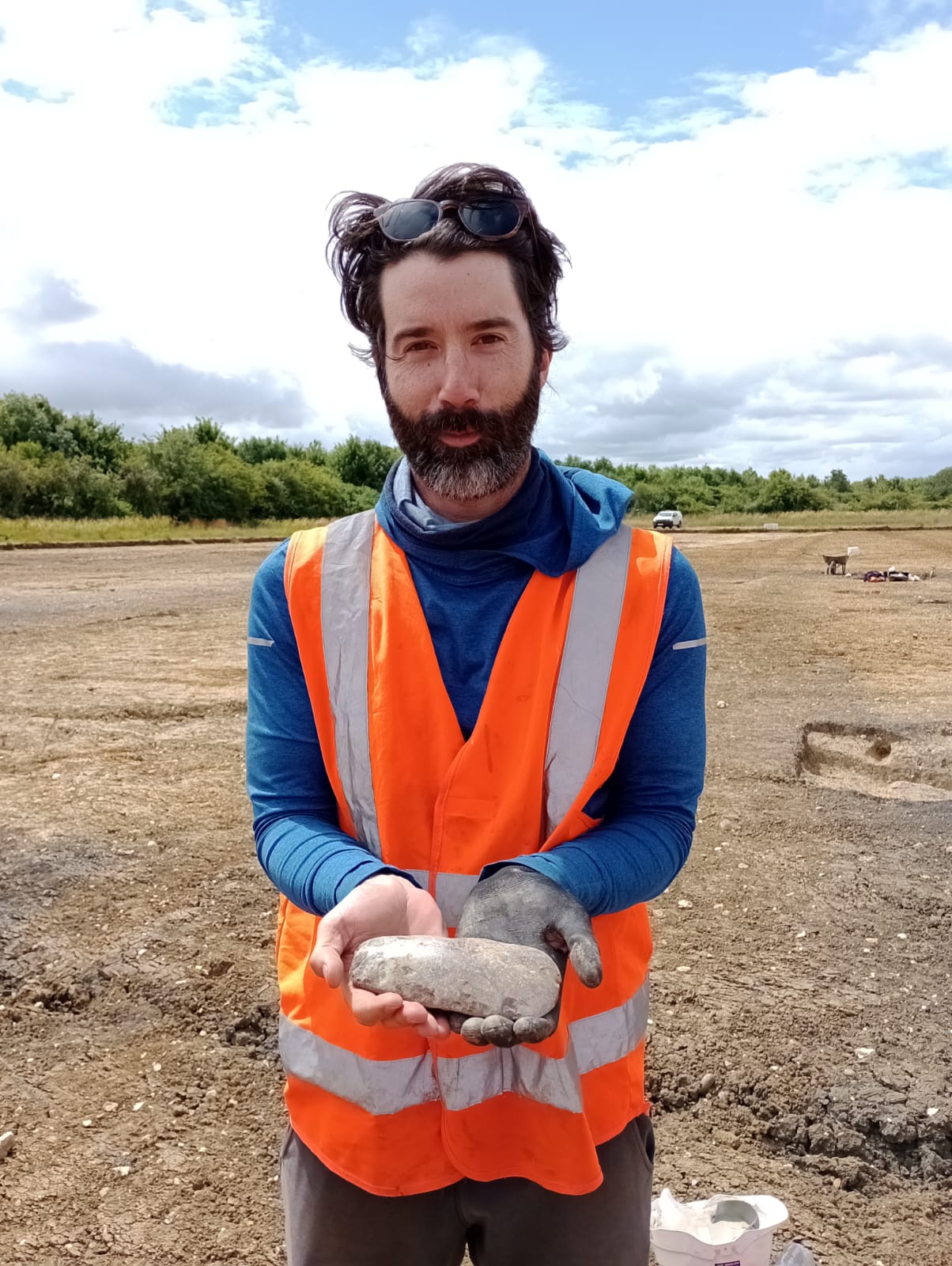 An archaeologist in an orange high vis vest holds a Neolithic axe head in a field stripped of top soil revealing archaeological features.