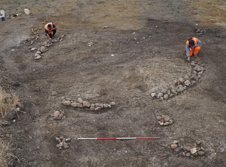 Two archaeologists in high visibility clothing are kneeling down in a field, using trowels to excavate a broken circular structure of stones, laid as a foundation for a Roman building.