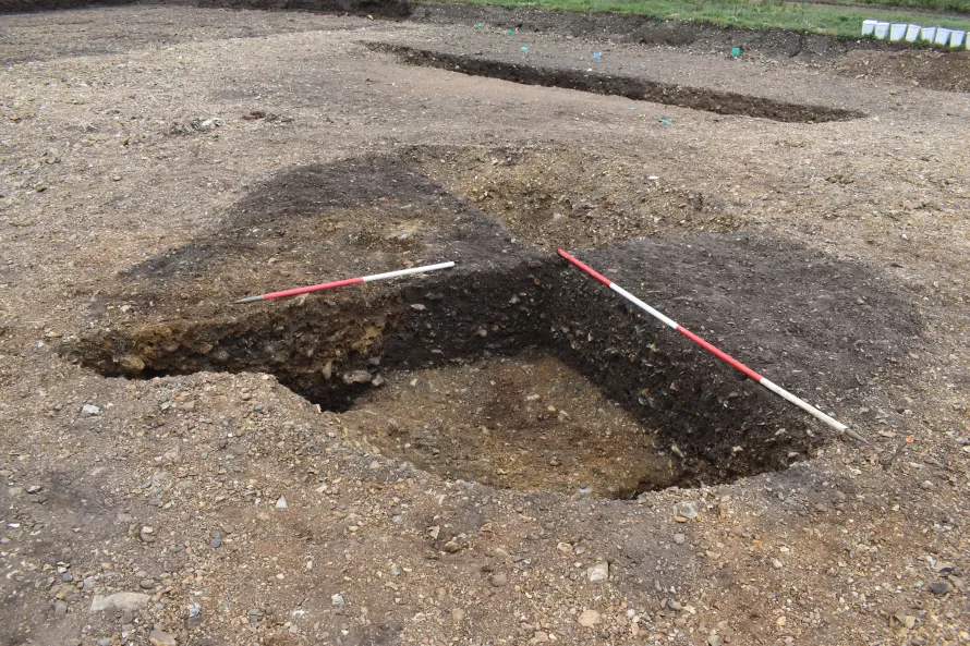A dark-brown sub circular feature is the in pale, stoney ground. Two opposing quarters have been excavated by archaeologists, red and white ranging poles sit on the edges ready for photographing.