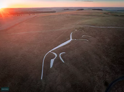 Aerial photo of the Uffington White Horse at sunrise, a prehistoric figure carved in the chalk of the downs. It is an abstract horse, with a long thin body, long tail. Its legs make it look as if it galloping over the hill. Image property of Hedley Thorne.