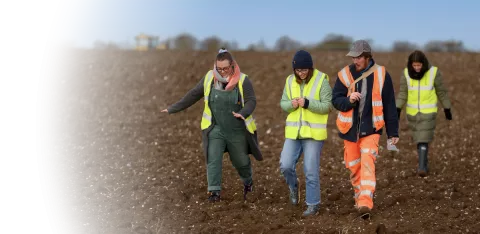 Archaeologist XX walks community visitors across a field whilst explaining and showing them finds from the project