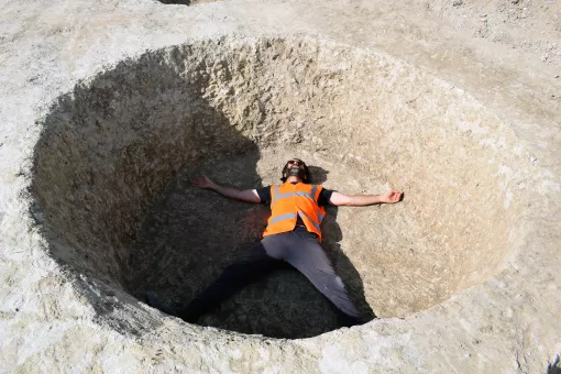 A male archaeologist lies down in a large circular pit with his arms and legs spread out
