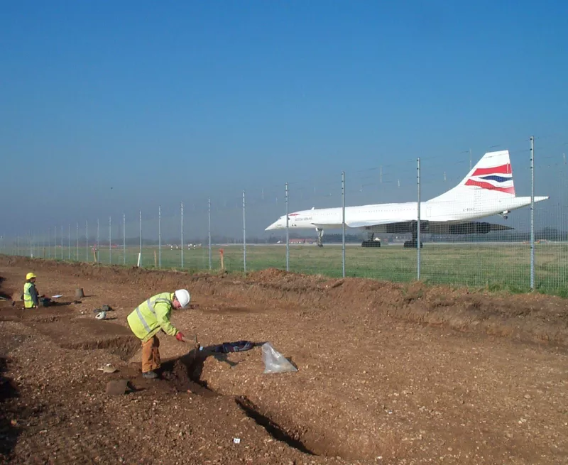 two archaeologists are digging near a fence that has a large commercial airplane on a tarmac