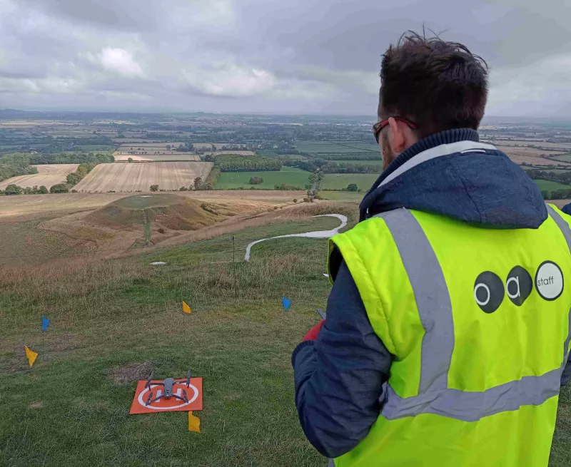 Man standing in the foreground with a remote control, ready to fly a drone over the white horse figure visible in the background