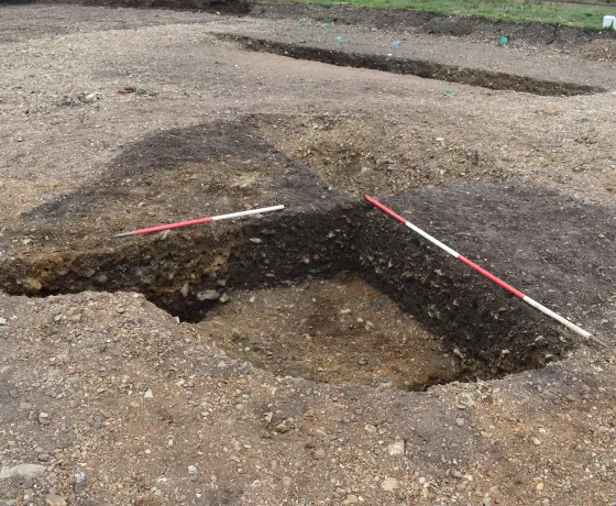 A dark-brown sub circular feature is the in pale, stoney ground. Two opposing quarters have been excavated by archaeologists, red and white ranging poles sit on the edges ready for photographing.